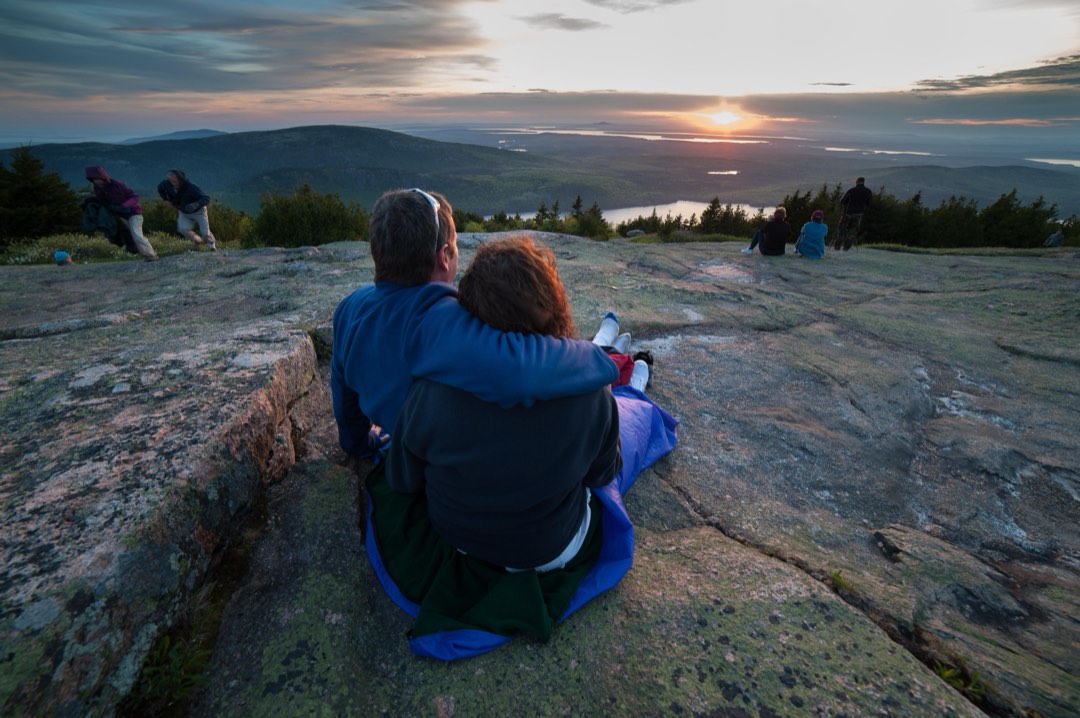 While seated on a blanket on a patch of mountaintop granite, a man and woman clasp their arms around