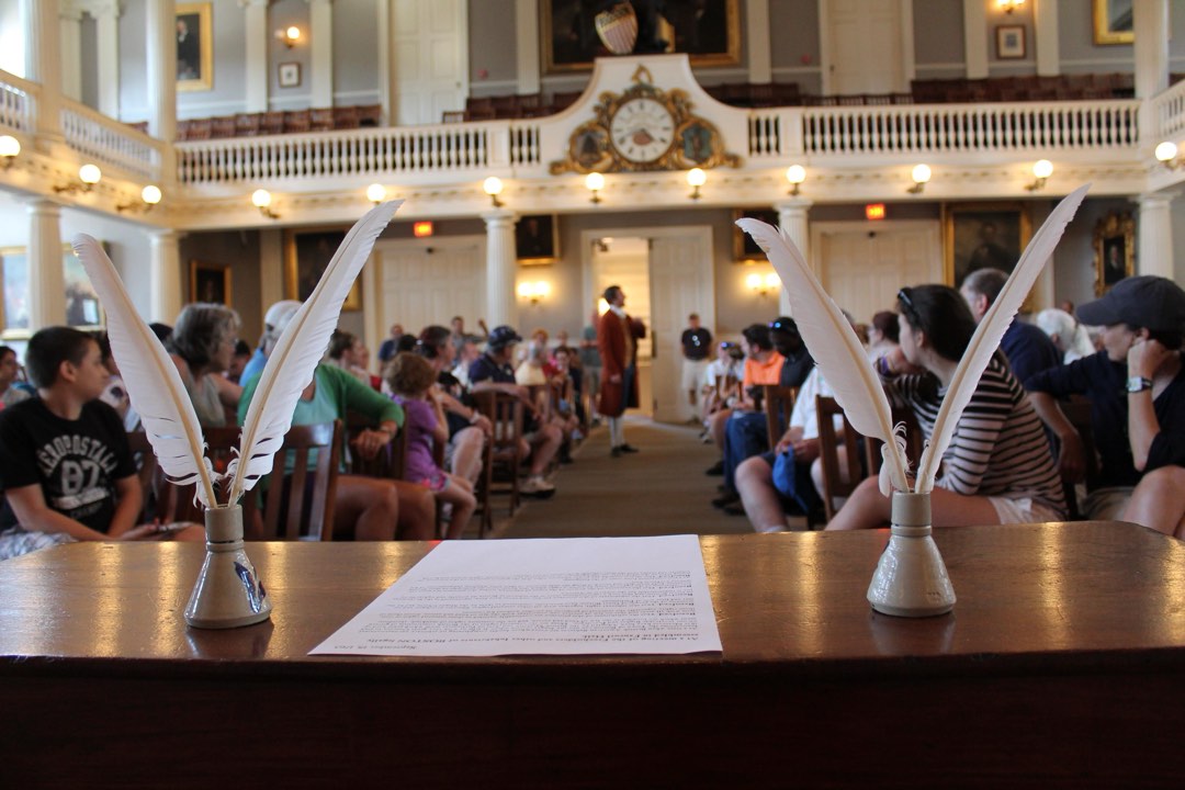 A desk with paper and two sets of quills frame a person in colonial wear standing in the aisle.