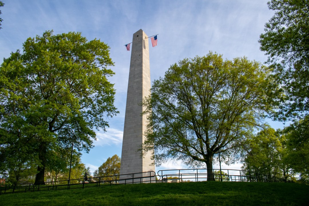 A tall white obelisk stands amongst leafy trees. Two colonial American flags hang from its windows