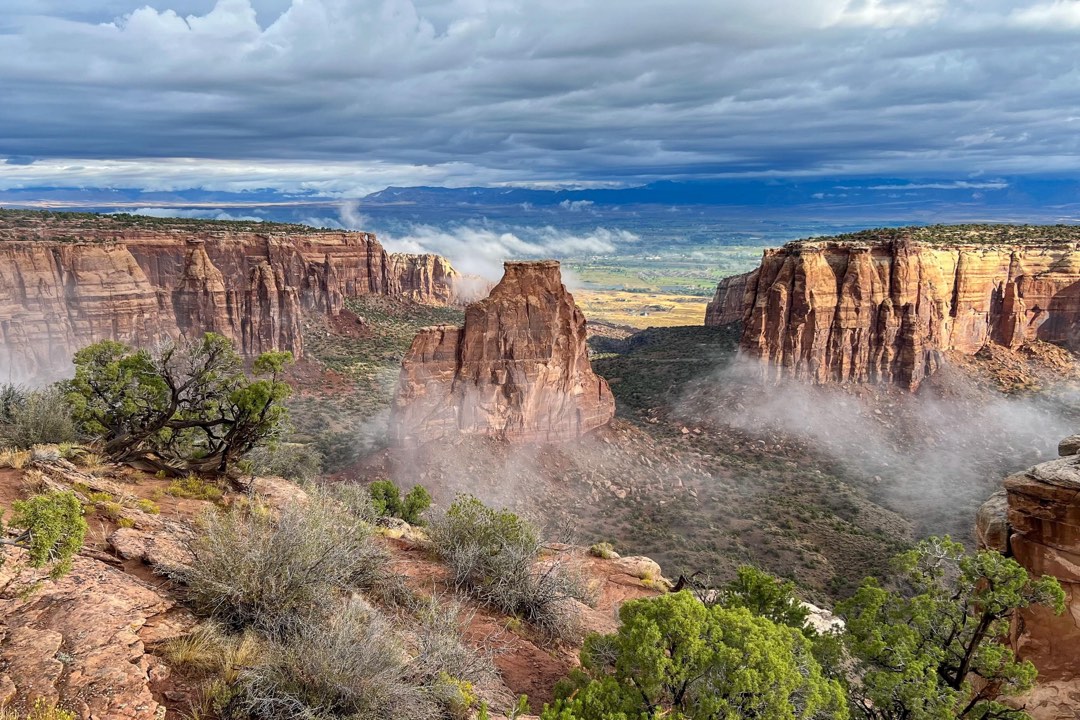 a wide sandstone monolith stands next to cliffs on a misty, cloudy day with green shrubs below