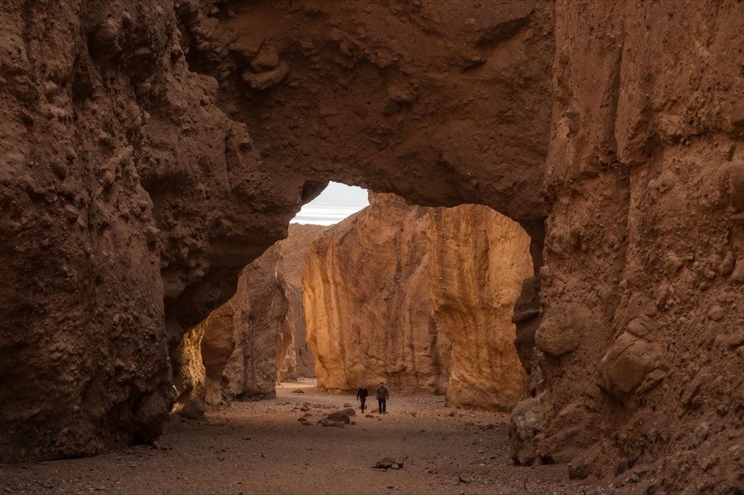 A natural rock arch spans a narrow desert canyon. The viewer stands in deep shade beneath the arch.