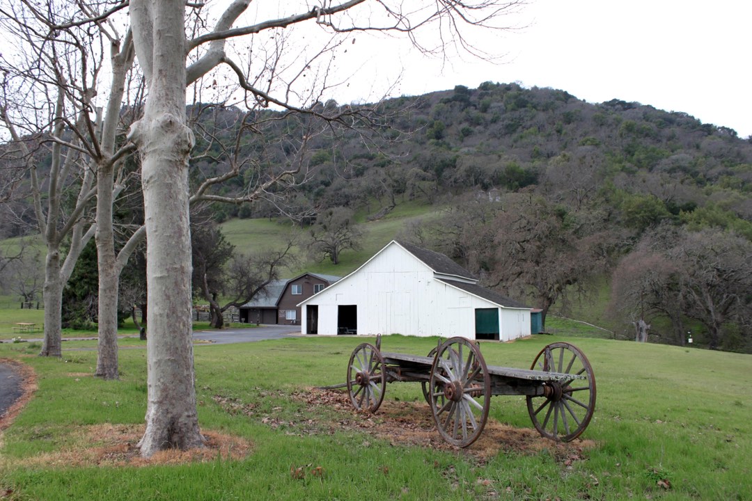An old barn and an historic wheeled wagon sit in front of a tree-lined hill.