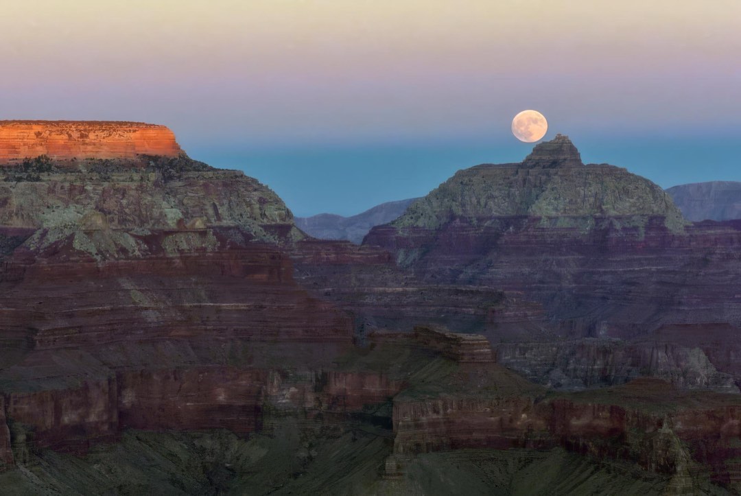 The last glow of sunset across the canyon rim with a full moon rising above a pointed peak