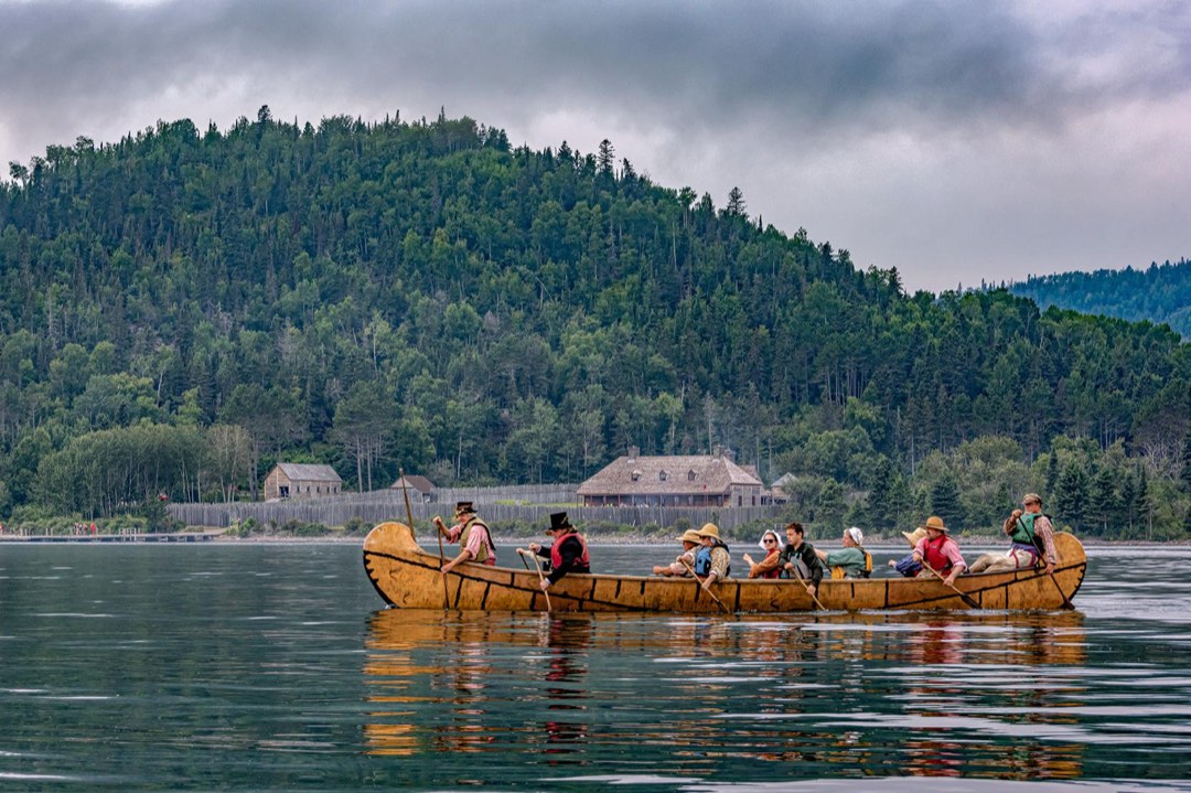 People in historic clothing paddle a canoe in a bay in front of a stockade.