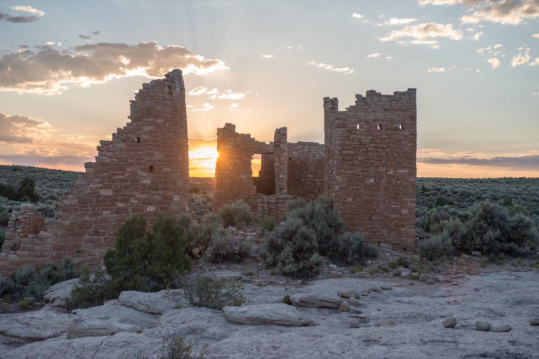 Tall stone structures with a setting sun shining through them. Desert landscape surrounds them.