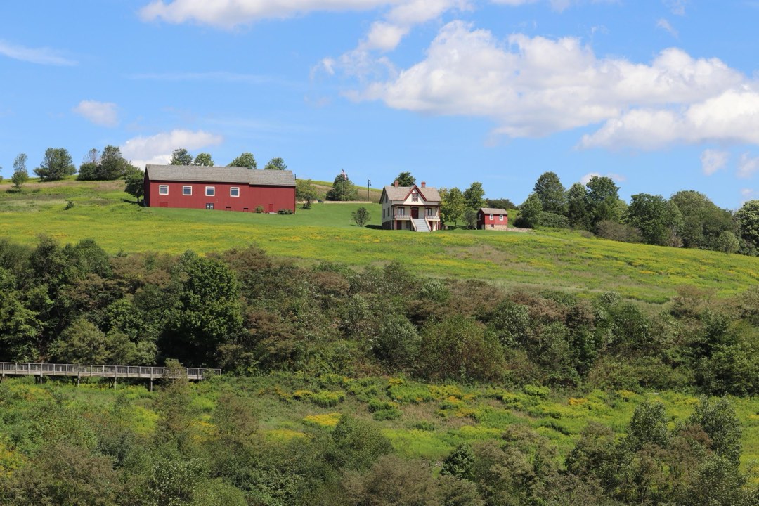 A farmhouse, shed, and long, red barn with windows atop a grassy hill