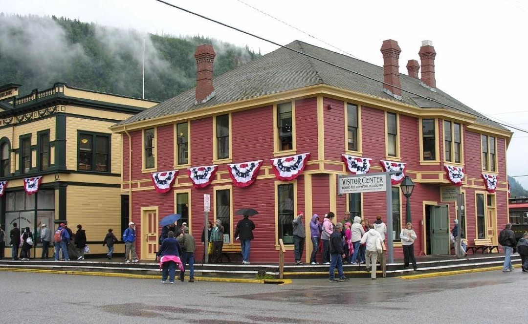 Visitors stand on a boardwalk outside a brick red colored wooden building with tidy golden trim.